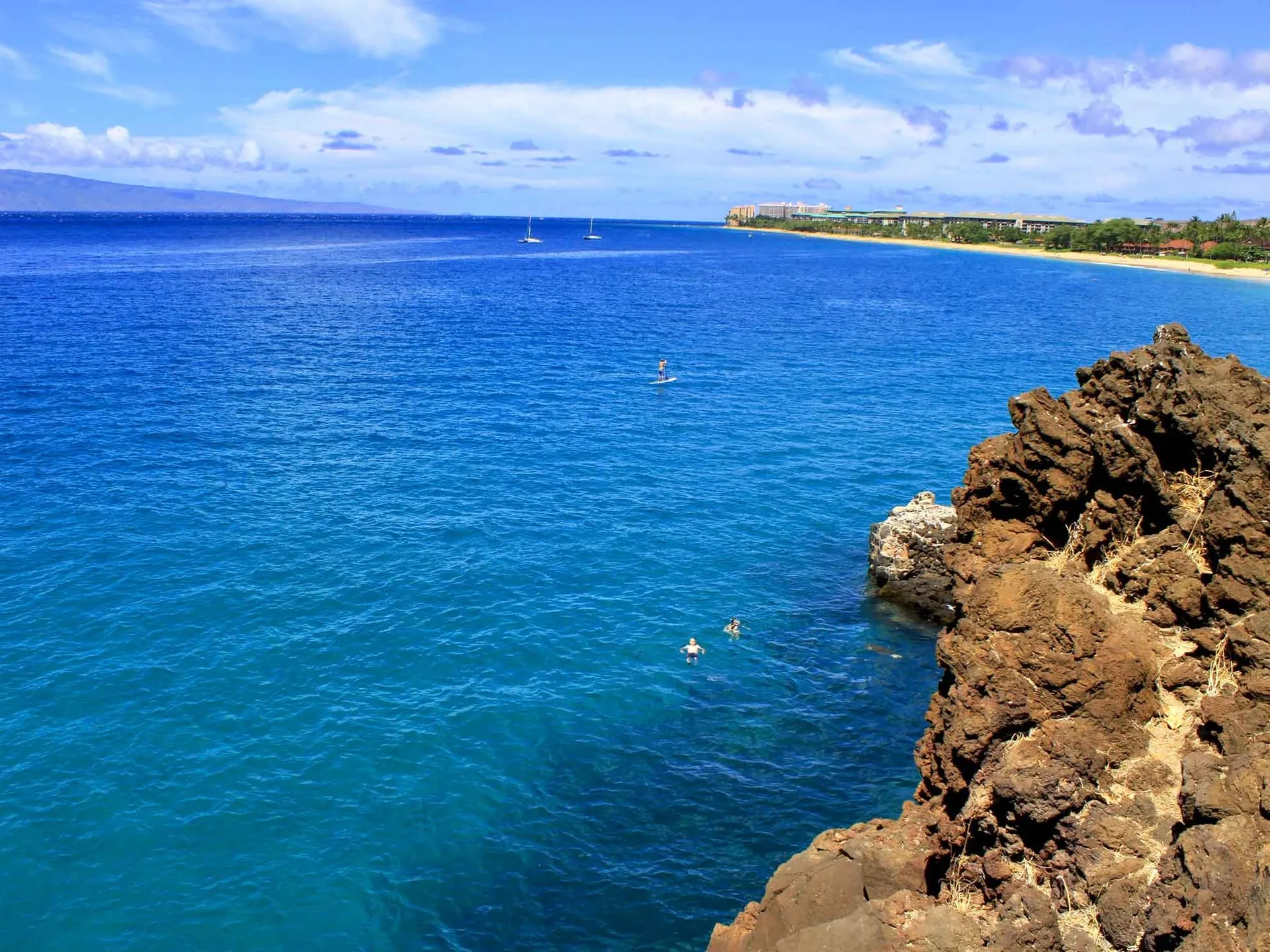 Snorkelers in clear blue water beside the lava of Black Rock at Kaanapali Beach, Maui.
