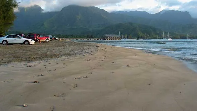 Sand, parked cars and the Hanalei pier at Black Pot Beach Park, with the mountains behind Hanalei Bay.