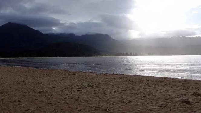 Sunlight breaking through rain clouds over Hanalei Bay, seen from the sand at Hanalei Beach Park Pavilion.