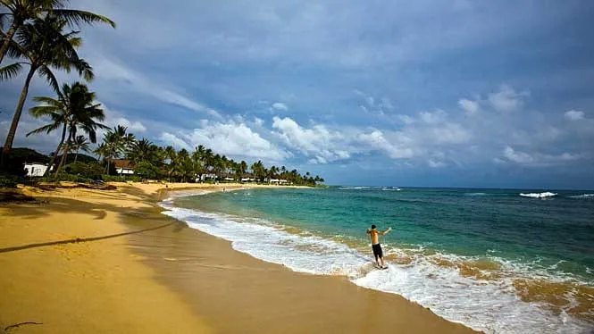 A fisherman casting from the sand at Poipu Beach, with palms and cottages behind.