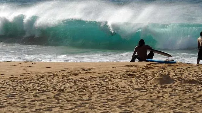 A bodyboarder sitting on the sand as a wave breaks at Sandy Beach.